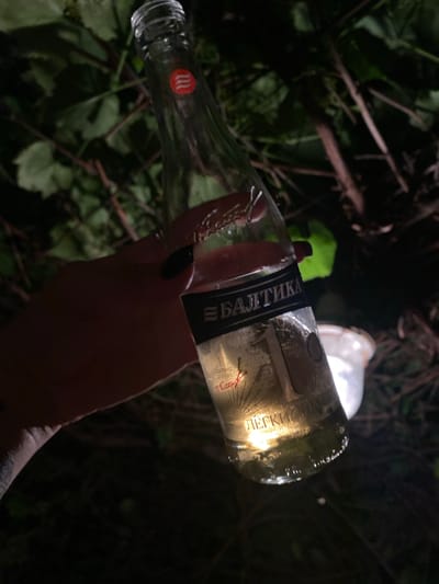 An extreme close-up, low-angle shot features a hand holding a clear glass bottle with a beverage. The bottle's label is primarily black with silver text, displaying Cyrillic lettering that reads "БАЛТИКА" and "ЛЕГКИЙ". The liquid inside is a pale yellow, and condensation is visible on the bottle. The background is dark and appears to be an outdoor setting with blurred foliage and branches, suggesting a natural environment possibly on the outskirts of Solnechnaya Polyana, Russia. The lighting is dim, with the bottle itself being illuminated from within and below. A white object, possibly a bucket, is visible in the background, out of focus.
