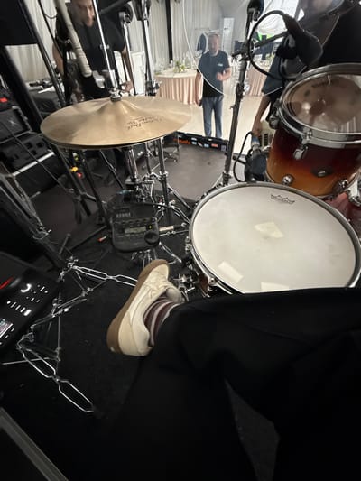 The image shows a drum set in what appears to be an indoor venue. The drum set includes a bass drum, snare drum, tom-toms, and a cymbal. A person's foot in a sneaker and striped sock is visible next to the drums. Cables, microphone stands, and other equipment are scattered around the drum set. In the background, a few people are visible, along with tables and decorations suggesting the setting is a performance or event space. The location is not discernible as Shchekutino, Russia, from the provided image.