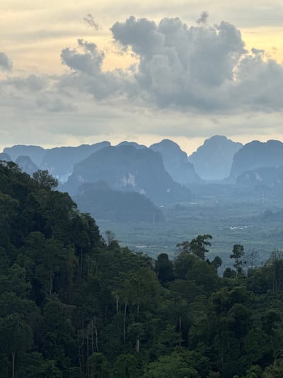The image captures a panoramic view of a mountainous landscape in the vicinity of Ban Khao Tang, Thailand. In the foreground, a dense, dark green forest covers a steep slope, with various trees of different heights and textures. The midground reveals a valley filled with more lush vegetation and scattered buildings, suggesting a rural settlement. Dominating the background is a series of majestic, mist-shrouded karst mountains, their distinctive shapes softened by atmospheric haze. The sky above is a canvas of soft, muted tones, with patches of pale yellow light breaking through a layer of dramatic, heavy grey clouds. The overall impression is one of natural grandeur and tranquility, with the interplay of light and shadow emphasizing the dramatic topography.