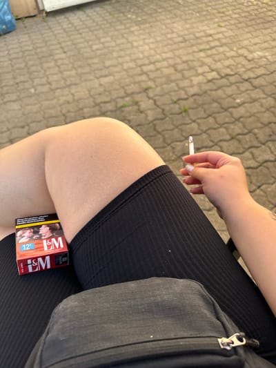 The image shows a person's legs and hand holding a cigarette, with a pack of L&M cigarettes on their thigh. The person is sitting outdoors on a paved area with interlocking stones. A black fanny pack is visible in the foreground. The background consists of a light-colored wall and some greenery. The overall setting appears to be a residential or public outdoor space. There is no visual information in the image that indicates the specific location of Bad Säckingen, Germany.