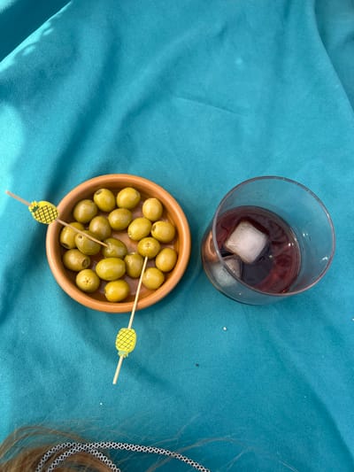 An overhead shot displays a wooden bowl filled with green olives and a glass of a dark red liquid with ice cubes, both resting on a textured teal fabric surface, possibly a tablecloth or blanket. Two cocktail picks with yellow pineapple decorations are inserted into the olives. The scene appears to be outdoors, with sunlight casting shadows on the fabric. Part of a person's hair and a patterned rope are visible in the lower section of the frame. This image does not provide any information about its location in Sant Pol de Mar, Spain.