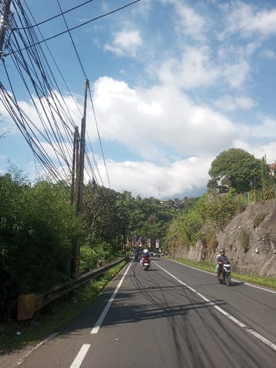 The image displays an ascending two-lane asphalt road in Ban Phonhxavanh, Laos, under a blue sky dotted with scattered white cumulus clouds. On the left side of the road, a metal guardrail runs parallel to a dense bank of green foliage, primarily bushes and trees. Above this vegetation, multiple utility poles support a complex network of black electrical and communication cables that extend across the upper left quadrant of the frame. In the distance on the left, a hillside covered with additional greenery features numerous residential structures and a prominent telecommunications tower. On the right side of the road, a grey concrete retaining wall with minimal vegetation is visible. The road itself has white solid and double-solid dividing lines. Several motorcycles are present on the road, all traveling away from the camera. In the foreground on the right, a person wearing a blue shirt and a black helmet operates a dark-colored motorcycle. Further down the road in the left lane, a red scooter carries at least one individual. Shadows from the overhead wires and poles are cast across the road surface.