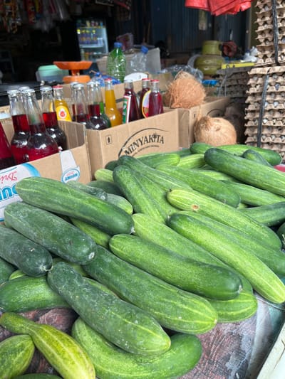 In Dayahbaro, Indonesia, a market stall displays a variety of produce and beverages. In the foreground, a large pile of fresh cucumbers is arranged on a table covered with a patterned cloth. Behind the cucumbers, several cardboard boxes contain glass bottles of red and yellow liquids. To the right, stacks of egg cartons are visible. In the background, a refrigerator unit is filled with drinks, and various other items and market structures are present. The scene suggests a typical outdoor market environment.