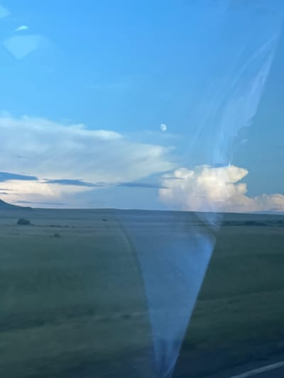 The media file captures a reflection, likely from a window, showing a blue sky with scattered clouds. A small, indistinct object, possibly a distant aircraft or balloon, is visible in the upper left portion of the sky. The reflection also includes a landscape scene, with hints of green fields and a darker, indistinct area that could be trees or a building. The overall impression is that of a bright, possibly sunny day, given the clarity of the blue sky. The orientation of the image is vertical, suggesting it might have been taken in landscape mode with the camera held sideways or that the reflection itself is distorted. There is no visible text in the media file.