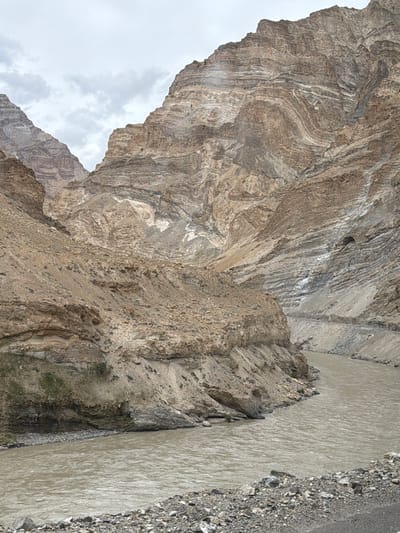 A wide, muddy river flows through a narrow gorge with steep, rocky mountains on either side. The mountains display distinct layers of sediment and rock, indicative of geological folding. The sky is overcast with clouds. The scene appears to be a natural landscape, likely in a mountainous region. There are no people or structures visible in the image. The overall impression is one of rugged natural beauty and geological history.