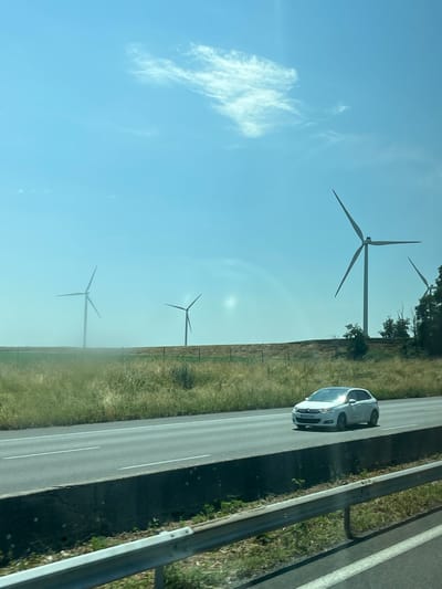 This media file depicts a scenic landscape featuring a road and wind turbines under a bright blue sky. The scene is captured from a moving vehicle, likely on a highway, as suggested by the perspective and the road markings. A white car is visible driving on the right side of the road.

In the background, several large wind turbines stand tall against the clear sky, indicating a renewable energy farm. The land adjacent to the road is covered in green grass and some vegetation, with a few trees visible near the wind turbines. The sun appears to be high in the sky, suggesting it is daytime, possibly midday or afternoon. The weather seems clear and pleasant, with a few wispy clouds adding texture to the sky.

The image is oriented vertically, with the sky taking up a significant portion of the frame. The tilt of the image suggests it might have been taken from a car window or dashboard, with the camera tilted upwards. There are no people clearly visible in the frame, but the presence of the car implies human activity. The overall mood is peaceful and evokes a sense of open space and natural beauty. No visible text is present in the image. The location context provided is Laucourt, France, which aligns with the presence of wind farms and rural road imagery typical of such areas.
