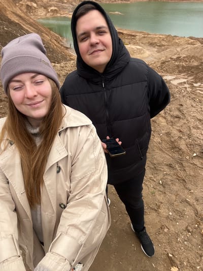 Two individuals are positioned close together in an outdoor setting. One person, dressed in a black hooded puffer jacket, black pants, and black sneakers, holds a smartphone. The second person wears a beige trench coat and a grey beanie. The background features a landscape of earth and water, possibly a reservoir or large body of water. The setting appears to be in Ivlevo, Russia.