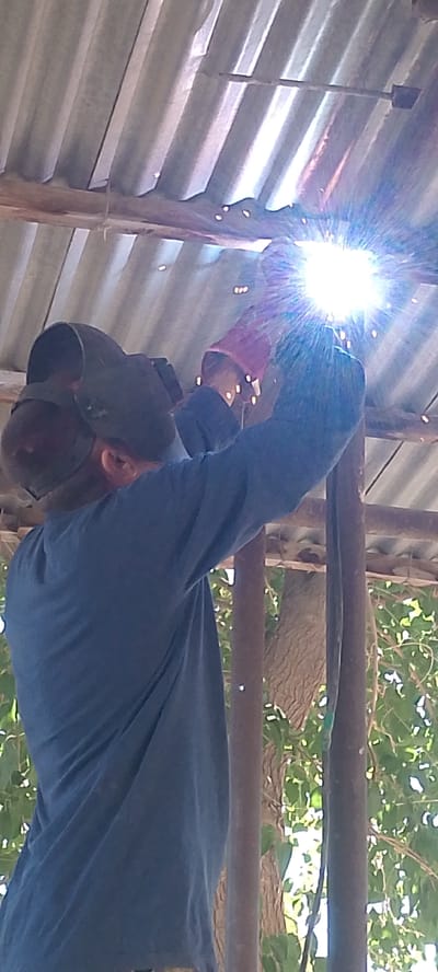 The image shows a person wearing a welding mask and gloves, actively welding under a corrugated metal roof supported by wooden beams. The welding process is producing a bright flash of light and sparks. The person is wearing a dark blue long-sleeved shirt. In the background, there are tree branches and leaves visible, suggesting an outdoor or semi-outdoor setting. The overall environment appears to be somewhat rustic. No specific location within Balkanabat, Turkmenistan is identifiable from the image content itself.