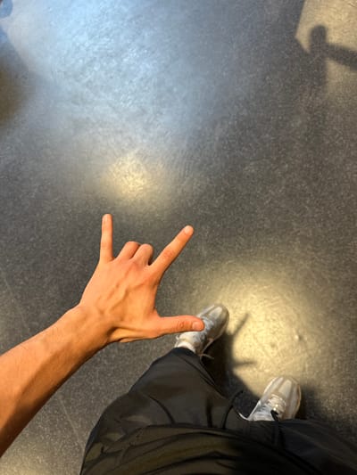 The image depicts a close-up, high-angle view of a person's left hand making a "rock on" gesture. The hand is illuminated, showing veins and skin texture. The person is wearing black pants and white sneakers. The background is a dark, textured floor, possibly asphalt or concrete, with faint shadows suggesting overhead lighting or a nearby structure. The scene is indoors, based on the artificial lighting and the floor material. There is no indication of the city of Alès, France, in the image itself.
