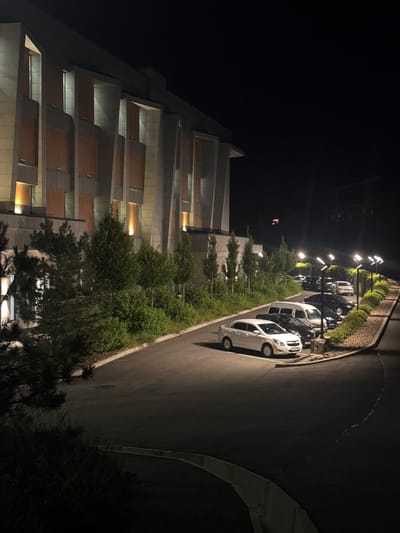 The image depicts a nighttime scene in Uchterek, Uzbekistan, featuring a modern, multi-story building with a unique architectural design characterized by sharp angles and alternating light and dark paneling. Numerous windows are illuminated from within, casting a warm glow. In front of the building, a paved road curves, lined with a row of streetlights. Several cars are parked along the roadside, including white sedans, black sedans, and a white van. Lush green trees and bushes are visible between the building and the parking area, with more dense foliage on the left side of the image. The overall impression is one of a well-maintained, contemporary establishment at night.