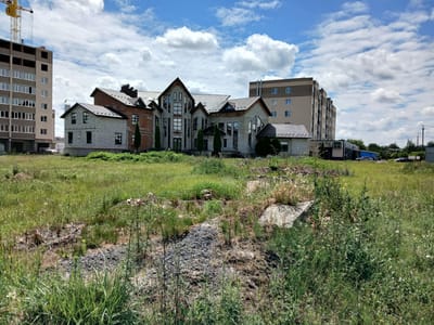 In Stryzhavka, Ukraine, a construction site is depicted under a partly cloudy sky. In the foreground, a field of tall green grass and weeds is visible, with scattered piles of gravel and debris. A large, light-colored concrete slab is present in the middle of the field. Dominating the background are several unfinished buildings. To the left, a tall, multi-story building is under construction, with a yellow construction crane positioned at its top. Adjacent to this, a distinctive building with a mix of red brick and light-colored brick facade, featuring multiple gables and large, angular windows, is partially completed. To the right of this, another multi-story building, appearing more finished with a beige and brown exterior, stands. Further to the right, a row of smaller buildings, possibly commercial, with dark facades are visible, along with several parked cars on a road. The scene suggests ongoing development in a semi-rural or suburban area.