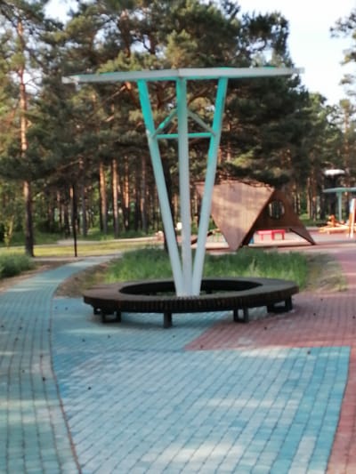 The image shows an outdoor public space with paved walkways and a seating area. In the foreground, a circular bench made of dark wood with visible cog-like detailing surrounds a white, three-legged structure with a horizontal beam at the top, illuminated with turquoise lights. The walkways are composed of light blue and red interlocking pavers. Behind the bench, a triangular wooden structure with a circular opening is visible, resembling a playhouse or shelter. The background features a dense forest of tall pine trees. This scene is located in Svirsk, Russia.