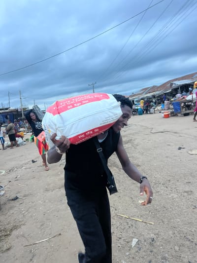 In Oko Amakom, Nigeria, a young man with dark skin, wearing a black sleeveless shirt and black pants, carries a large white sack of rice labeled "Pure Refined Thai Parboiled Rice, 50 Kgs Net" over his left shoulder. He is walking to the right of the frame and smiling. A black strap from a bag is visible across his chest. In the background, a woman with dark skin and a colorful skirt is also carrying a sack over her shoulder, walking to the left. The environment appears to be a busy market with stalls and people in the background. The ground is dirt and debris. The sky is overcast.