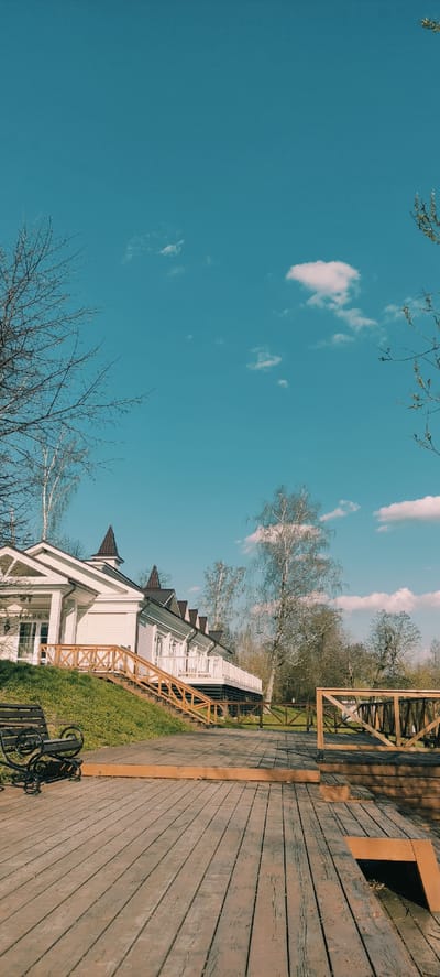 The image captures a scene in Grebnevo, Russia. A white, two-story building with a brown roof and decorative gables stands on a grassy lawn. The building is partially obscured by leafless trees in the foreground. A wooden boardwalk with a brown railing extends to the right of the building, leading out of frame. The sky is a clear blue with scattered white clouds. A portion of a bicycle is visible near the building.