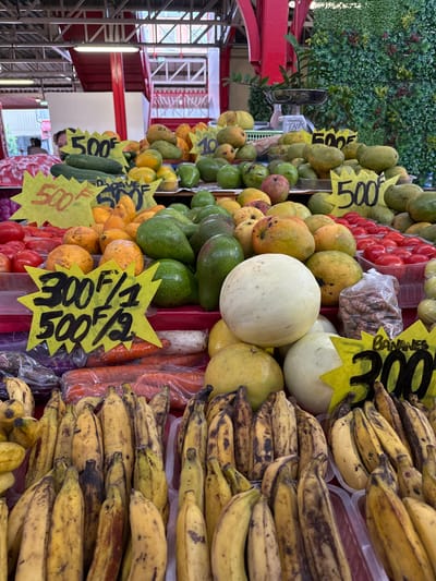 The image displays a bustling fruit and vegetable stall within an indoor market, specifically Le Marché de Papeete (Papeete Public Market) in Papeete, French Polynesia. The scene is dominated by tiered displays of fresh produce under an overhead metal structural ceiling with red beams.

In the foreground, numerous bunches of ripe, mostly yellow and brown-speckled bananas are arranged in plastic trays. A yellow star-shaped price tag reads "BANANES 300F." To the left, a similar yellow sign states "300F/1 500F/2" above clear plastic-wrapped carrots. Various melons are present, including pale yellow-green and off-white spherical types.

The midground features abundant piles of green avocados, intermingled with ripe mangoes exhibiting yellow, orange, and reddish hues. Bright red tomatoes are neatly presented in clear plastic containers on the right side. Other tropical fruits like papayas and more mangoes are visible on higher tiers. Additional yellow star-shaped price tags, reading "500F" and "500F/2," are interspersed among the produce. A small plastic bag of what appears to be dried nuts or legumes is situated behind the melons on the right.

In the upper background, a silver weighing scale is visible against a green wall of artificial foliage. To the far left, a blurry figure of a person is partially visible behind a red and white patterned umbrella. The overall environment is an active market setting with a wide variety of fresh, colorful produce.