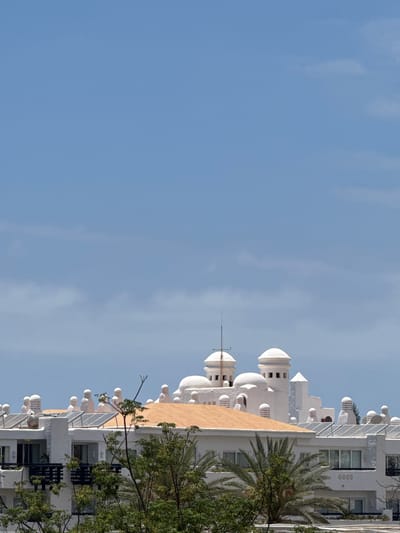 The image shows the distinctive white, domed architecture of buildings in San Eugenio Alto, Spain, under a clear blue sky with wisps of white clouds. The structures feature multiple rounded domes and minaret-like towers of varying sizes, some with dark-capped windows. A prominent orangish-brown tiled roof is visible on a lower section of the building. Solar panels are installed on some of the flat roof areas. Lush green trees and palm fronds partially obscure the lower levels of the buildings, suggesting a warm, sunny climate. The foreground is dominated by foliage, while the background consists of the sky and the upper portions of the distinctive buildings. No people are visible in the frame.