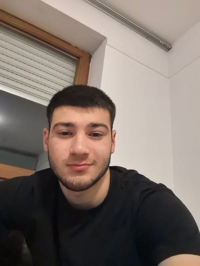 A close-up photograph depicts a young man with dark hair and a short beard wearing a black t-shirt. He is indoors, likely in a room with white walls. A window with closed blinds is visible behind him. The location is not discernible as Aystetten, Germany based on the visual information in the image.