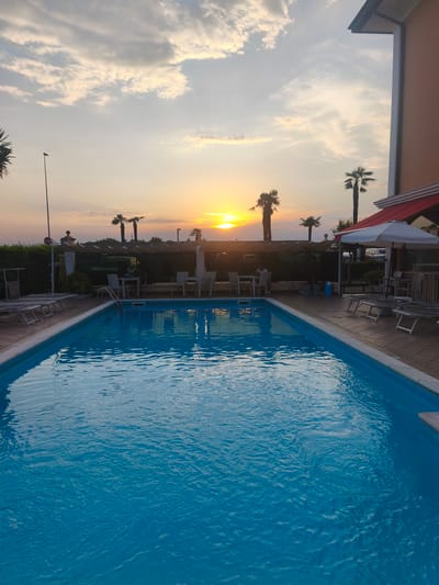 The image depicts a swimming pool area at sunset, with the sun low on the horizon and casting a warm glow across the sky. The pool is rectangular and filled with clear blue water, reflecting the sky and surrounding objects. Several lounge chairs are arranged on the tiled patio surrounding the pool, some of which are empty. A few palm trees are visible in the background, adding to the tropical ambiance. A building, likely part of a hotel or resort, is situated to the right of the frame, with a red awning over an outdoor seating area. The sky is partly cloudy, with streaks of orange and yellow from the setting sun. The scene appears to be located in Albignasego, Italy, given the provided context. There are no people visible in the pool itself, but one person is seated at a table in the background area, under a covered patio. The overall impression is one of a tranquil evening setting.