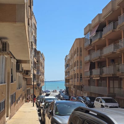 The image depicts a street in Guardamar Del Segura, Spain, with tall, multi-story buildings on either side. The buildings are beige and have balconies. Cars are parked along the right side of the street. A group of people walks on the sidewalk between the cars and the buildings. At the end of the street, the blue sky and ocean are visible.