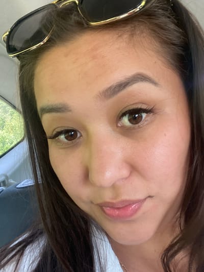 A close-up, overhead selfie of a woman wearing sunglasses, taken from a low angle, captures her face from the eyebrows down. She has long, dark hair that frames her face, and her skin appears smooth with subtle makeup. The background shows a blurry view of green trees and what appears to be the interior of a vehicle. The scene is set outdoors, possibly in a park or a suburban area.