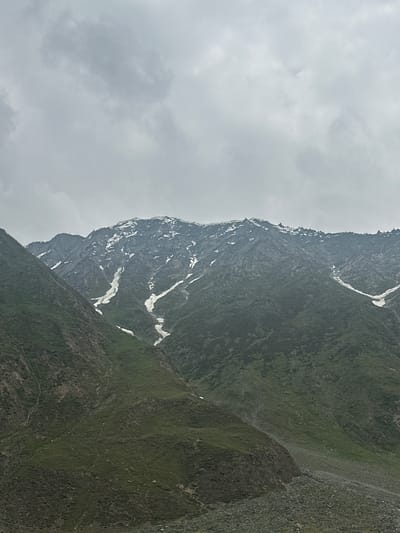 The image shows a cloudy sky over a mountainous landscape in Matayan, India. The mountains are covered in green vegetation and patches of snow, with rocky slopes visible in the foreground. The sky is overcast with grey clouds, indicating a potentially cool or damp day. The scene is a natural, outdoor environment with no visible signs of human activity or structures.