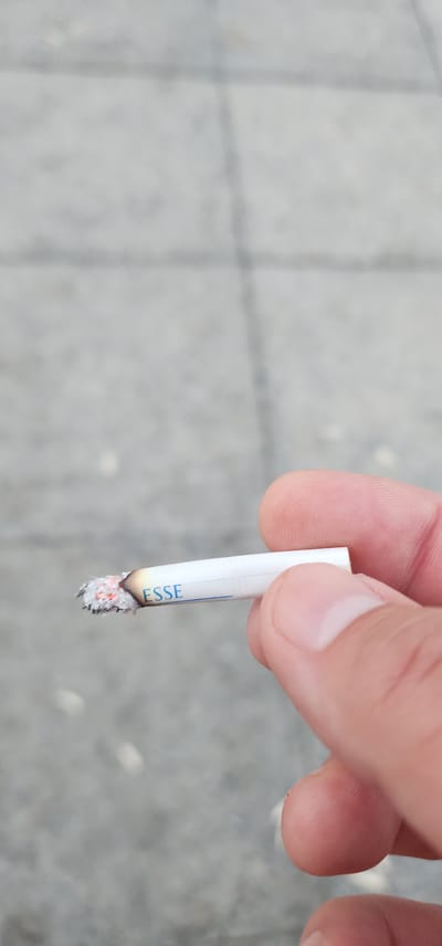 In Türkmenbaşy, Turkmenistan, a person's fingers hold a lit cigarette with the brand name "ESSE" visible on its white filter. The cigarette is smoldering at the tip, with ash and glowing embers present. The background is a blurred, gray surface that appears to be pavement or a similar textured outdoor ground. The lighting suggests it is daytime.