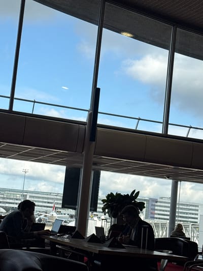 The image displays an interior view of a lounge or terminal at Paris Charles de Gaulle Airport in Tremblay-en-France, France, looking outwards through a large window wall. The upper section of the image shows a blue sky with scattered white clouds, with a faint, elongated white object visible in the upper left. Below the window line, an airport tarmac is visible, featuring a commercial aircraft with a red stripe on its fuselage and multiple multi-story airport buildings in the background.
In the foreground, a long, light-colored table occupies the mid-ground. Two adult males are seated at this table, largely silhouetted. The male on the left, dressed in dark clothing, is looking at an open laptop screen while bringing a cup to his mouth. The male to his right, wearing a dark jacket and glasses, is leaning forward, seemingly focused on papers or a device on the table. A dark green potted plant is positioned on the table between them. A third individual is partially visible in the right background, seated with their back to the viewer. The interior lighting is subdued compared to the bright daylight outside, creating strong contrasts and shadows on the foreground subjects.