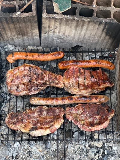 This overhead shot captures a rustic outdoor barbecue scene, likely set in Mirkovo, Bulgaria. The primary subjects are various meats cooking on a metal grill grate positioned over hot embers and ash.
The grill holds three thick cuts of meat, resembling pork steaks or chops, alongside three sausages. The steaks exhibit distinct char lines and a browned, cooked surface, suggesting they are nearing completion or have been flipped. Two of the sausages are a reddish-brown hue, indicating a seasoned or cured variety, while the third is lighter in color. All the items appear to be actively grilling, with some surfaces glistening from rendered fat.
The grill setup itself is makeshift, constructed from rough, grey hollow concrete blocks visible on the left side of the frame. These blocks show signs of soot and wear from previous use. Beneath the grill grate, a mixture of grey ash and visible red embers provides the heat for cooking. To the far left, a small fragment of green material and a thin wooden stick are present, resting against the concrete structure.
The bright, even lighting suggests the photograph was taken during the daytime. There are no people visible in the image, nor is there any discernible text. The scene evokes a casual, outdoor culinary activity.