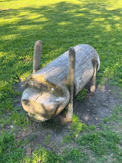 The image shows a wooden sculpture of a pig in a park. The sculpture is made from a log and has two wooden posts for legs at the front and two at the back. It is positioned on a grassy area with some gravelly patches. The lighting suggests it is daytime. This sculpture is located at Tuiranpuisto park in Oulu, Finland.