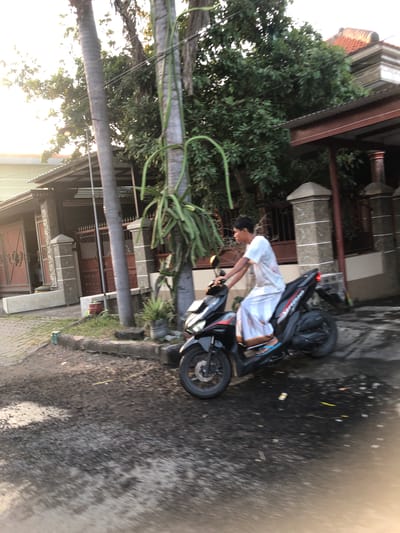 A young man wearing a white robe and blue flip-flops rides a black scooter on a paved road. The man is looking to his left. The scooter has a yellow turn signal on the front and a red taillight on the back. The road is wet and reflects the sunlight. There are palm trees and houses in the background. The houses have red tile roofs. The scene appears to be in the city of Sentul, Indonesia.