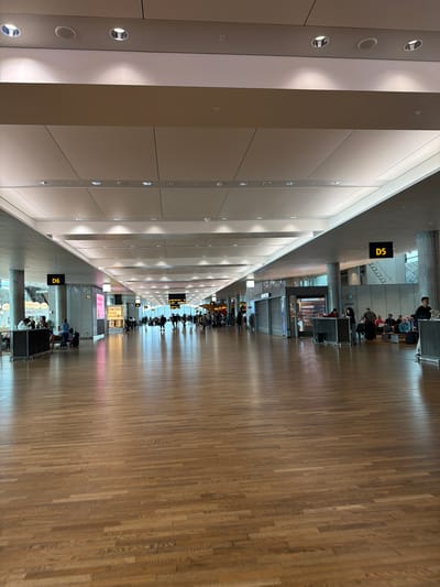 This is an image of the interior of Oslo Airport, Gardermoen, Norway. The scene depicts a wide, modern airport terminal hallway with a polished wooden floor that reflects the overhead lighting. People are visible in the distance, walking and seated in waiting areas. There are shops and kiosks along the sides of the hallway. Overhead signs indicate gate numbers, with "D6" visible on the left and "D5" on the right. The architecture is contemporary, featuring a high ceiling with integrated lighting and a neutral color palette. The overall atmosphere is one of a busy but orderly transportation hub.