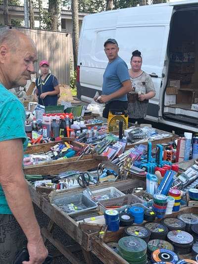 In the city of Belaya Kholunitsa, Russia, an outdoor market is in full swing. In the foreground, a man with a bald head and a turquoise shirt stands with his back to the camera, facing a stall laden with tools and supplies. The stall displays a variety of items, including bottles of liquids, spray cans, tools, electrical tape, and cutting discs.
Behind the stall, a white van is parked, its rear doors open, revealing stacked boxes. Three individuals are visible near the van. A man in a blue t-shirt and a baseball cap stands next to the van, appearing to be a vendor or customer. To his right, a woman with dark hair tied up in a bun, wearing a grey t-shirt, holds some cash. Further to the left, a woman in a navy blue vest with yellow trim and a pink cap is also present. The market setting is suggested by the presence of the stalls and the casual outdoor environment with trees in the background. The overall atmosphere appears to be that of a bustling local marketplace.