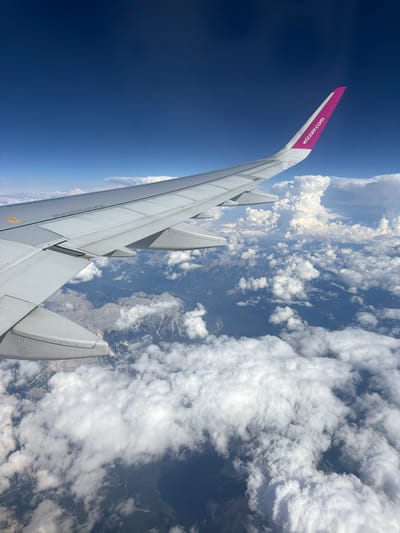The image, taken from the window of an airplane, shows a view of the earth below and the sky above. The airplane's wing, a part of the Wizz Air aircraft, is prominently visible in the foreground, extending across the frame. The wing is white with a vibrant pink tip, featuring the "wizzair.com" logo.
Below the wing, the landscape is a mix of rugged mountains, appearing grey and sparsely vegetated, and a dense covering of white, fluffy clouds. The clouds create a textured layer, partially obscuring the terrain beneath. Glimpses of dark, possibly shadowed areas, can be seen through gaps in the clouds, suggesting valleys or darker landforms. The sky above is a deep, clear blue.
The scene depicts an aerial view, likely during daylight hours, with sunlight illuminating the clouds and the top of the wing. There is no indication of the city of Leno, Italy, or any specific identifiable landmarks within this aerial perspective. The image focuses on the airplane's wing and the natural environment seen from altitude.