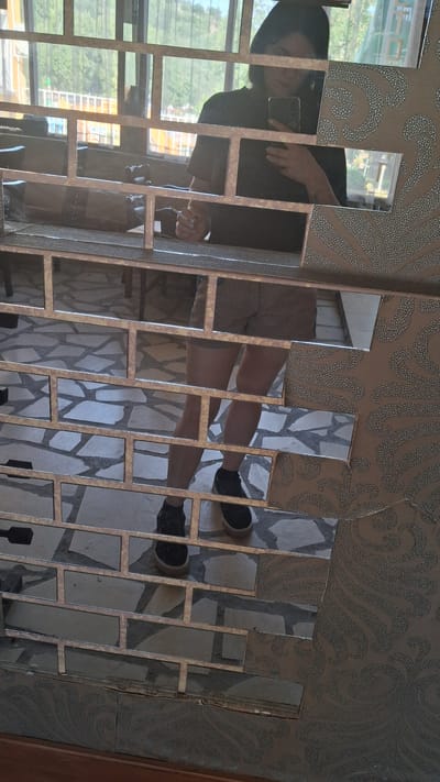 In Marchevo, Bulgaria, a person is photographed in a room with a tiled floor and textured wallpaper. The person, wearing shorts and black sneakers, is holding a smartphone, seemingly taking a selfie. The foreground features a decorative mirror mosaic, which partially obscures the person and their surroundings. The mirror is framed by a textured surface, likely part of the room's decor. The background shows a window with trees and buildings visible outside.