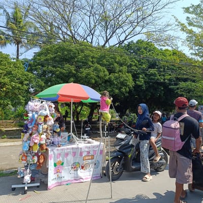 In the city of Kebondalem, Indonesia, a street vendor is selling plush toys under a colorful umbrella. The toys are displayed on a rack that is positioned in front of a table covered with a printed cloth. Behind the vendor's stall, a monkey dressed in a pink jacket and green pants is perched on a pole, seemingly part of a performance or display. Several people are present, including a woman in a blue hijab and a young girl on a motorcycle, as well as other individuals walking nearby. The scene takes place outdoors on a sunny day, with trees and a clear sky in the background. The overall atmosphere appears to be that of a bustling local market or roadside stall.