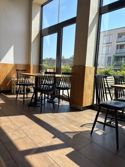 The image depicts the interior of a modern café with large windows overlooking an outdoor area. The café has a minimalist aesthetic with wooden paneling on the walls and a tiled floor. Several black chairs with wooden frames are arranged around wooden tables, suggesting a seating area for patrons. The sunlight streams in through the windows, casting distinct shadows on the floor and walls. Outside, a contemporary building with balconies and windows is visible, along with green foliage and trees, indicating an urban or suburban setting. The sky is clear and blue. The scene appears quiet and unoccupied, with no people present. The style of architecture and the presence of a modern building suggest that this café is likely located in Elsenfeld, Germany, a town known for its modern developments.