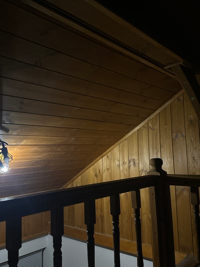 The image depicts an interior view looking upwards at a wooden sloped ceiling and wall. The ceiling is constructed of horizontal wooden planks, while the adjacent wall is made of vertical wooden planks. A dark wooden railing with turned balusters is in the foreground, suggesting the scene is located on a staircase or balcony. A contemporary light fixture with a geometric metal cage is visible in the upper left corner, illuminating the wooden surfaces. The overall impression is of a rustic or chalet-style interior, likely residential. The location is identified as Brestnik, Bulgaria.