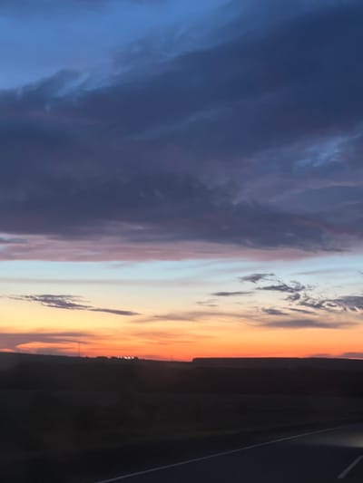 The image depicts a sunset over a rural landscape, likely in the vicinity of Bagrash-Bigra, Russia. The sky is filled with a gradient of colors, transitioning from deep blue and purple at the top to orange and yellow near the horizon. Clouds are present in various shades of gray and purple, some appearing wispy and others more dense. Below the sky, a dark silhouette of rolling hills or a flat plain stretches across the frame. In the distance, lights from a town or settlement are visible along the horizon. The foreground shows a dark, paved surface, possibly a road, with a white line marking its edge. The overall impression is one of a serene, natural landscape at dusk.