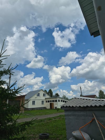 A tall, green conifer tree branch is in the foreground, obscuring the left side of the image. Beyond the tree, a rural scene unfolds under a partly cloudy blue sky. A pale yellow, two-story house with a gray metal roof is visible, featuring multiple windows. To the left of the house is a wooden structure with a darker garage door. In front of the house, a dirt road passes through a grassy area. To the right of the house, a long, single-story building with a row of windows is attached. In the immediate foreground, a gray cinder block structure with a corrugated metal roof is present, with a rake and two bags leaning against it. A single tire lies in the grass to the left. In the background, other houses and trees are visible. The image appears to be taken in Sokur, Russia.
