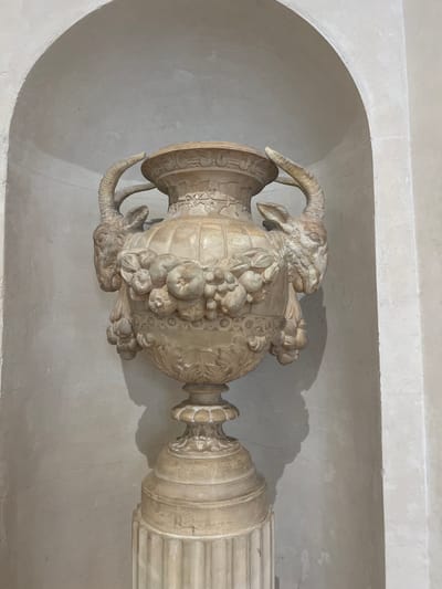 An ornate, light-colored stone vase with ram's heads and fruit carvings sits on a fluted pedestal within a recessed, arched alcove. The vase's handles curl upwards and outwards, seamlessly transitioning into the carved heads of rams, whose horns curve back. A garland of fruits, possibly apples and grapes, adorns the vase's body, with smaller clusters hanging below. The alcove's interior walls are textured and slightly uneven, with subtle linear details along the top curve. The exterior wall of the alcove is a smooth, light surface. The scene is lit from above and to the right, casting a soft shadow of the vase onto the back wall of the alcove. This vase is located in the Espace Liberté in Évian-les-Bains, France.