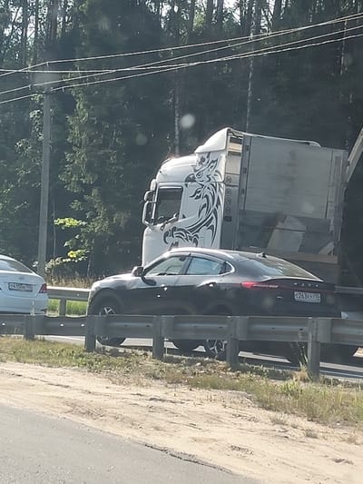 In this image, a dark-colored sedan is on a highway, partially obscured by a large white truck. The sedan's rear is visible, displaying a license plate that reads "E263KY 790". The truck has a distinctive tribal-like graphic on its side, resembling a stylized bird or animal. The scene appears to be on a sunny day, with lush green trees lining the background. A guardrail separates the highway from a grassy and sandy shoulder. Another white sedan is partially visible in the far left of the image, with its license plate showing "T415CB 790". The overall setting suggests a rural or highway environment in Russia, possibly during daylight hours given the bright illumination and shadows. No specific activity or event is depicted, other than vehicles traveling on a road. The image is taken from a slightly elevated perspective, looking down at the vehicles and the road.