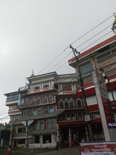 The photograph shows a multi-story building with ornate architectural details, including arched windows, decorative trim, and a rooftop dome. To the right is a red and white structure, possibly a petrol station, with associated signage. Power lines intersect the view, creating a web against the overcast sky. People are visible at the base of the building.