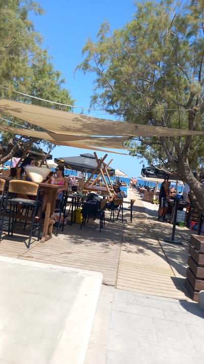 This is a tilted, eye-level shot of a beachside cafe or restaurant in Tigaki, Greece. The scene is outdoors, under a clear blue sky. Several tables and chairs are set up on a wooden deck, some covered by beige sunshades and umbrellas, suggesting a sunny day.

In the foreground, a woman in a pink bikini and shorts is seated at a table, holding a drink. Various bags, including a yellow one, are visible near the tables. In the background, more tables and chairs are arranged closer to the beach, where many people are seen lounging and swimming in the sea. Trees with green foliage provide shade over the seating area.

Notable details include the presence of Coca-Cola branding on a black umbrella. The atmosphere appears relaxed and leisurely, typical of a beach resort on a warm day, likely midday given the strong sunlight and shadows. The image composition and subject matter suggest a vacation or a day at the beach.