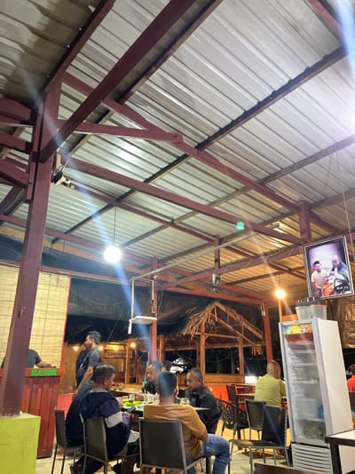 The image captures a casual indoor or covered outdoor eating establishment, likely a restaurant or cafe, at nighttime. The perspective is tilted counter-clockwise, looking up towards the ceiling and across the dining area.

The prominent feature of the structure is its roof, which is made of corrugated metal sheets. This roof is supported by an intricate framework of dark red-painted metal beams and natural wooden rafters, creating a rustic industrial aesthetic. Several bare light bulbs or floodlights are suspended from the ceiling, illuminating the space and creating noticeable lens flares due to their brightness against the dark background. A small ceiling fan is also visible, hanging from one of the beams.

Hanging from the ceiling structure on the right side of the frame is a framed picture, showcasing two men. One appears to be wearing a white turban or head covering, and the other is in a lighter-colored outfit, suggesting a portrait of significant individuals. Below this picture, a tall, white commercial display refrigerator, likely stocked with beverages, is visible.

Below the refrigerator, several men are seated around tables, engaging in what appears to be a social gathering. They are dressed casually, and some have drinks on their tables. Their faces are not clearly discernible due to the distance, angle, and lighting. In the background, beyond the immediate seating area, more of the establishment's structure can be seen, including some natural thatched materials used in the construction, suggesting an open-air or semi-open-air design.

In the bottom right corner of the image, a green box-like structure is visible with the word "KASIR" clearly written on it, indicating a cashier station. Further in the background, a faint sign on a wall or structure reads "D'WOPS KITCHEN" and potentially "CAFE" below it, identifying the establishment. The artificial lighting, the darkness outside the illuminated areas, and the social gathering suggest the time of day is evening or night.