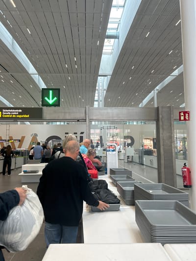 The image shows a security checkpoint at an airport. It is located inside a modern building with a high ceiling and a grid of white lighting. The checkpoint is set up with gray bins and trays, and a metal detector. There are several people in line waiting to pass through the checkpoint. A man with a black jacket is holding a tray, and a woman with a red jacket is standing behind him. A green arrow sign above the metal detector is pointing downwards. Several people are walking through the checkpoint. The airport is located in the city of Granadilla, Spain.  The coordinates provided (latitude: 28.047, longitude: -16.58) do not correspond to the location in the image. The image appears to be from an airport in Europe.  The image is in a neutral color palette of whites, grays, and blacks. The overall impression is of a busy airport with security measures in place. The image was likely taken with a digital camera at a slight angle.
