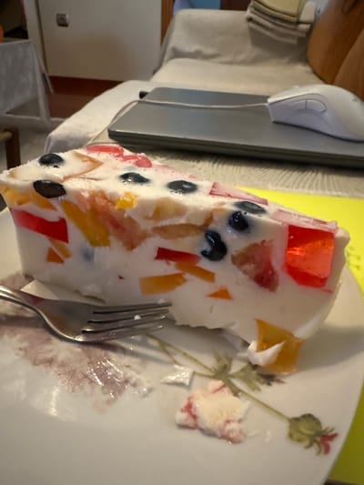 A slice of fruit gelatin dessert, containing pieces of red, orange, and yellow gelatin, as well as blueberries, sits on a white plate with a floral pattern. A fork rests on the plate, slightly to the left of the dessert. The dessert appears to have been partially eaten, with some crumbs and stray pieces on the plate. In the background, out of focus, is a grey laptop with a white computer mouse and a white cable. Behind that, a light-colored couch or chair is visible, and a white cabinet door with a black knob. The scene appears to be indoors, likely on a table. There is no indication of the city of Ilion, Greece, or any specific geographic location.