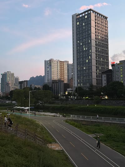 The image depicts a view of a cityscape in Uijeongbu-si, South Korea. In the foreground, a paved pathway curves through a grassy area with trees and what appears to be outdoor exercise equipment. Several people are visible on the pathway and near the equipment, some walking or standing, and others seemingly engaged in exercise. The pathway has distinct yellow lines marking lanes, suggesting it is a designated route for cycling or walking.
Behind the pathway and green space, a row of tall, modern apartment buildings dominates the midground. These buildings vary in architectural style and color, with many windows illuminated, indicating it is likely evening. In the distance, a mountain range with a distinctive, jagged peak is visible, silhouetted against a soft, pale sky with streaks of pinkish clouds, characteristic of either sunrise or sunset.
Further to the left, additional buildings are visible, including one with visible Korean text. One sign appears to say "800-1776" in white text on a black background. Another sign has red Korean characters. The overall scene suggests a peaceful urban environment blending nature with residential and recreational spaces. The lighting indicates it is either dusk or dawn.