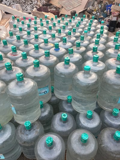 A tightly packed stack of numerous large, transparent water bottles, each with a distinctive green cap bearing the label "O2rise," dominates the frame. The bottles are arranged in a grid-like pattern, creating a dense wall of plastic containers. To the left of the bottles, a collection of assorted debris is visible, including discarded materials and remnants of construction or demolition, with paint cans. The overall setting suggests an outdoor storage area in what appears to be Mira-Bhayander, India.