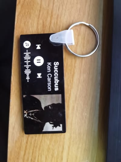 A black rectangular keychain, approximately 8 cm in length, is depicted resting on a light brown wooden surface with visible grain, within the city of Dolno Izvorovo, Bulgaria. A metallic split key ring is attached to the upper edge of the keychain via a translucent plastic connector. The keychain's face displays a music player interface design. In the upper left quadrant, a musical note icon is visible. Along the left side, a vertical pattern of varying length lines resembles a sound waveform or equalizer. Central to the design are playback control icons: a rewind arrow, a pause symbol (two vertical bars), and a fast-forward arrow. Vertically oriented along the right side of the keychain, the text "Succubus" and "Ken Carson" is displayed, likely indicating a song title and artist. Below this text, a dark, silhouetted image of a person's profile is visible, with faint details suggesting chains or necklaces around the neck. To the right of the wooden surface, a dark, vertically textured object or surface is partially visible.