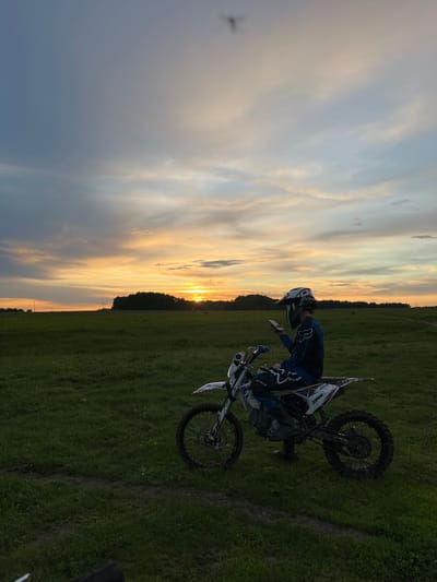 A person in motocross gear, including a helmet and blue and black clothing, is standing next to a dirt bike on a grassy field. The dirt bike is mostly white with black accents. The sky behind them is a gradient of colors, transitioning from a dark blue/grey on the left to a bright orange at the horizon, suggesting it is either sunrise or sunset. A line of trees is visible on the horizon. The location is Новочувашский, Russia.